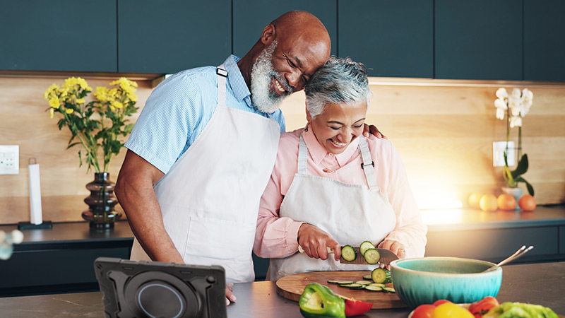 Smiling senior man and woman preparing fresh vegetables in kitchen using his Lingraphica TouchTalk AAC device.
