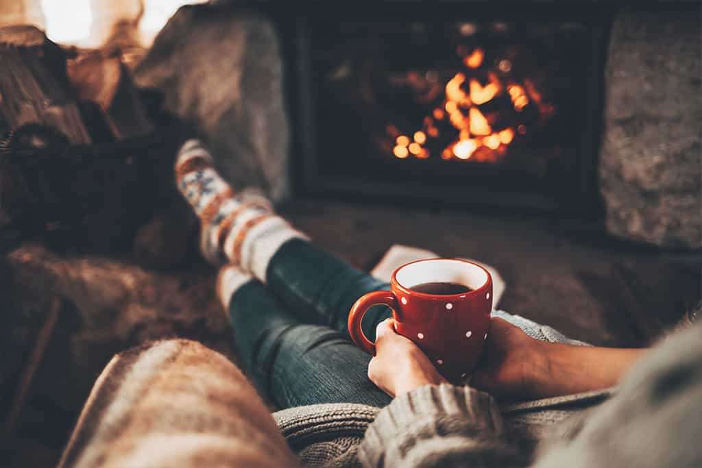 Person sitting by a lit fireplace holding a red mug of tea and wearing cozy wool socks