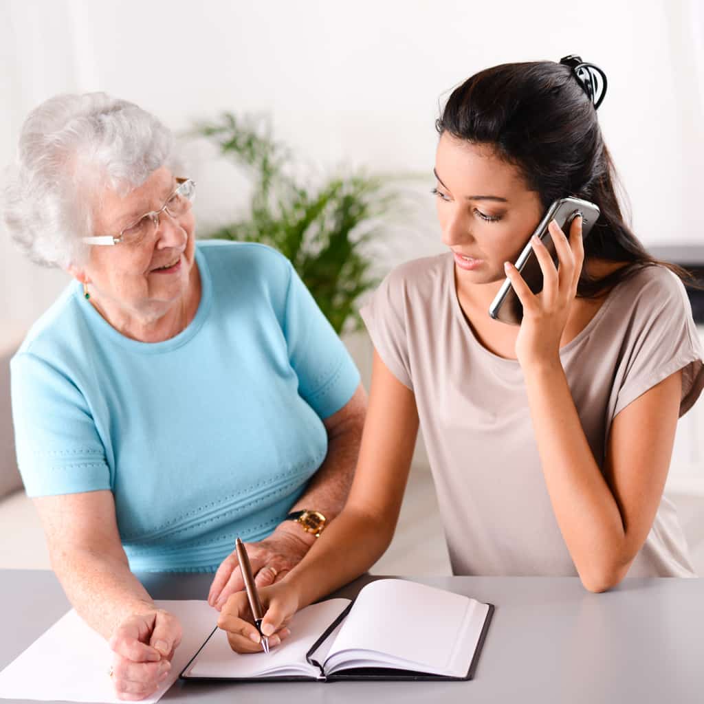Caregiver talking on phone with elderly love one looking on.