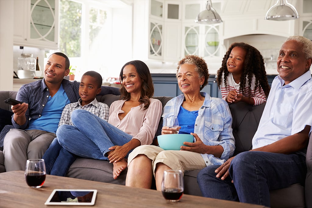 Multi-generational family sitting on the couch enjoying a television show, drinking and eating snacks